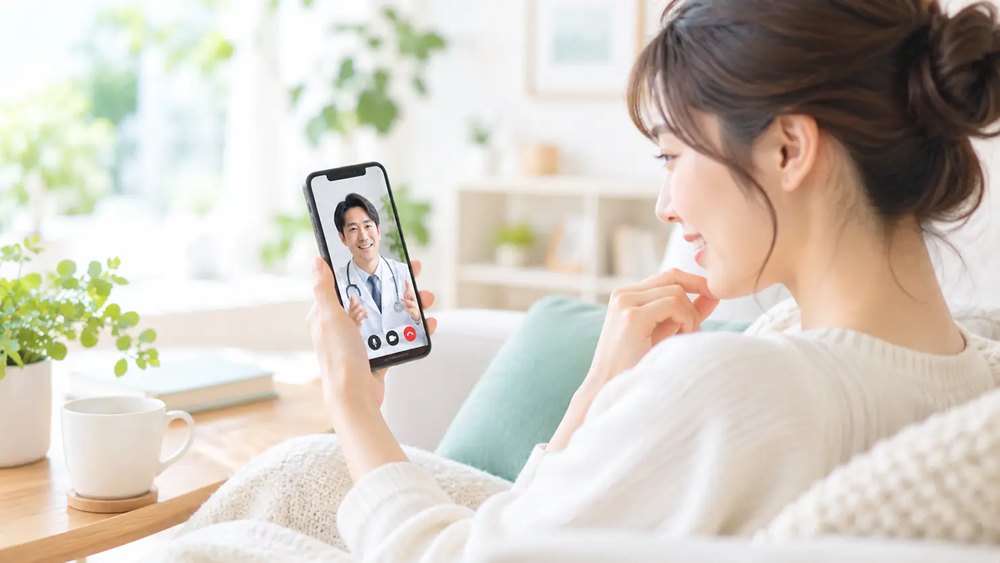 自宅のソファでスマートフォンを使い、医師とビデオ通話でオンライン診療を受ける女性